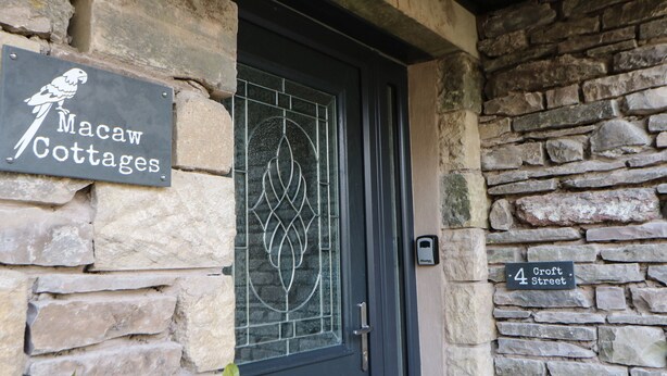 A stone cottage entrance with a dark door, a window with a decorative pattern, and signs indicating 'Macaw Cottages' and '4 Croft Street'. - Macaw Cottages No 4 (Kirkby Stephen)