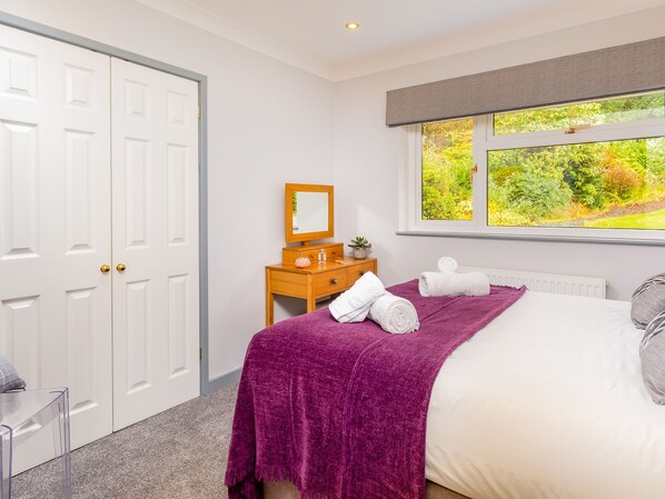 A bedroom with a bed, a window with a view of greenery, a wooden dresser, and a white door.