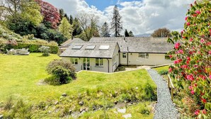 Une maison moderne avec un toit en pente, une terrasse dotée de mobilier d’extérieur et un jardin bien entretenu, agrémenté de diverses plantes et d’un chemin de gravier.