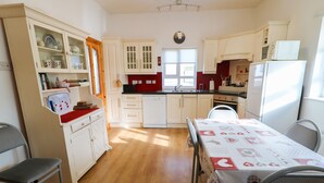 A kitchen with white cabinets, a dining table with a red and white tablecloth, and a window with a view of greenery outside. - Ard an Phíobaire (Bunbeg)