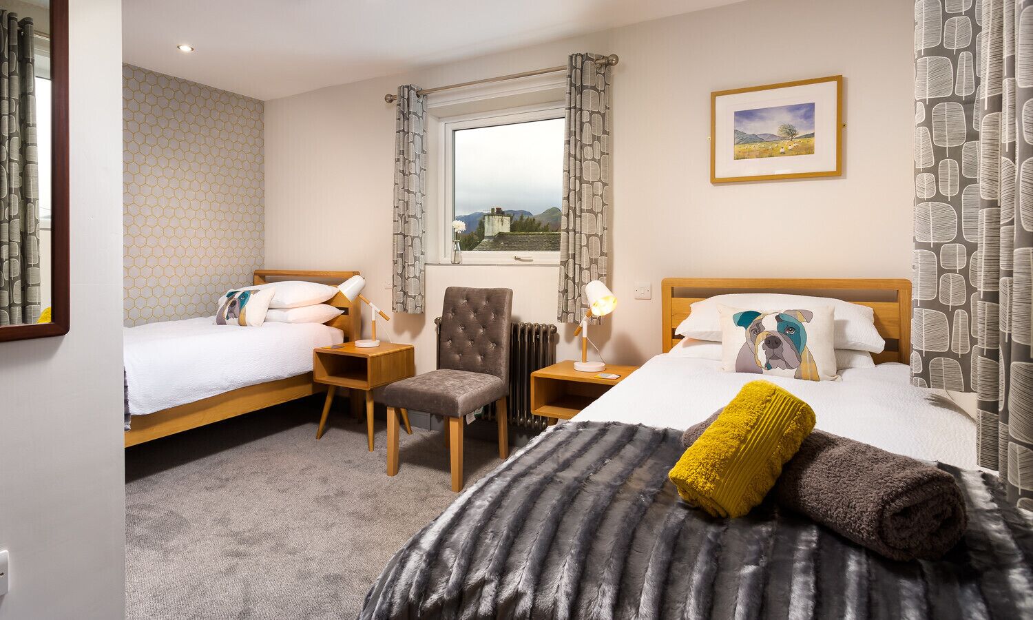A hotel room with two single beds, a chair, a nightstand with a lamp, a window with curtains, and a framed picture on the wall.