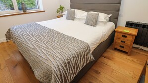 Un lit bien fait, avec une tête de lit grise, une table de chevet dotée de tiroirs, un radiateur et une fenêtre donnant sur un paysage verdoyant.