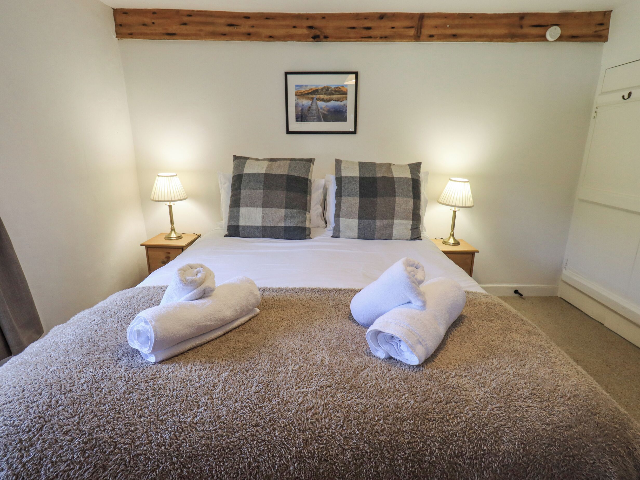 A neatly made bed with two pillows, two bedside lamps, and two rolled towels on a textured carpet.