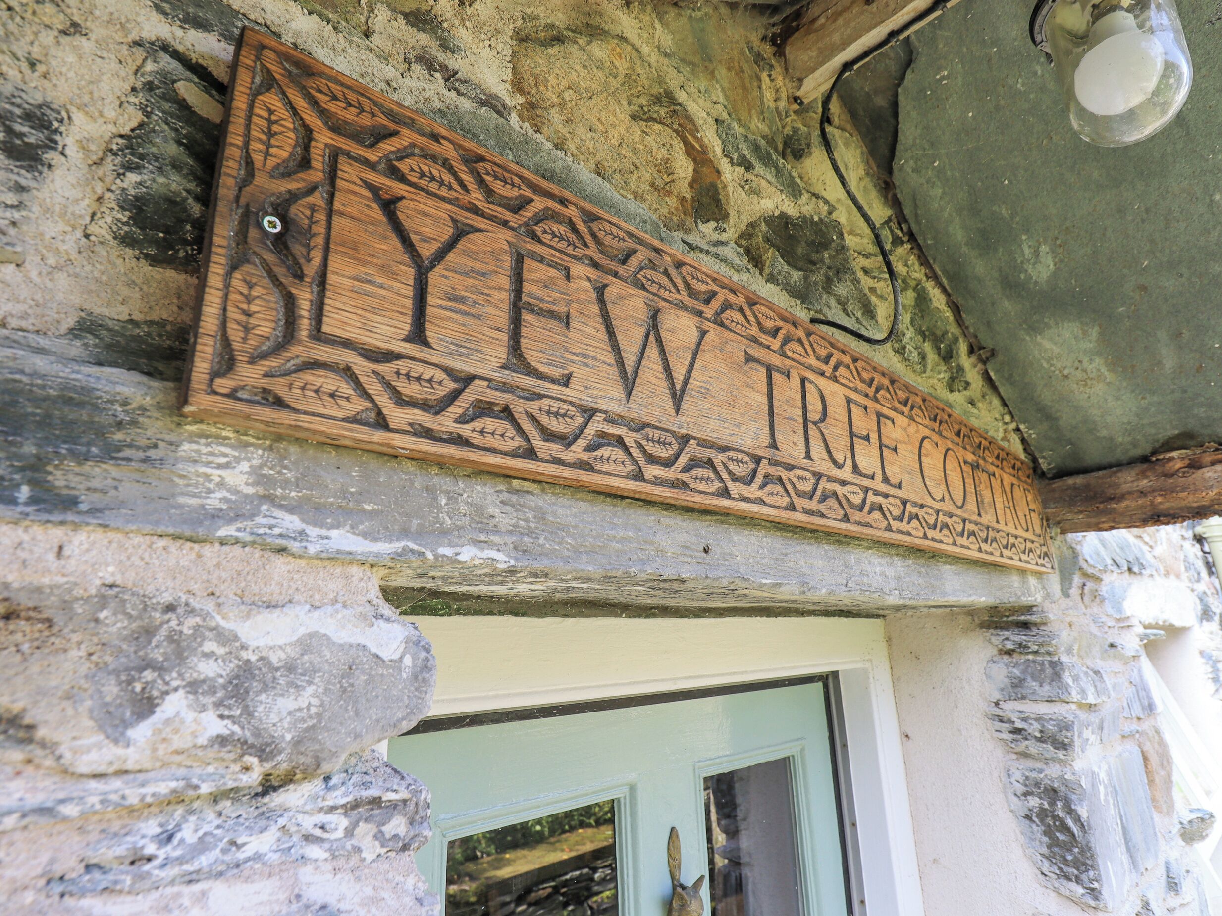 A wooden sign with intricate carvings and the text "LYEW TREE COTTAGE" mounted on a stone wall.