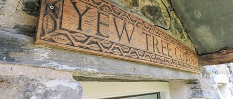 A wooden sign with intricate carvings and the text "LYEW TREE COTTAGE" mounted on a stone wall.