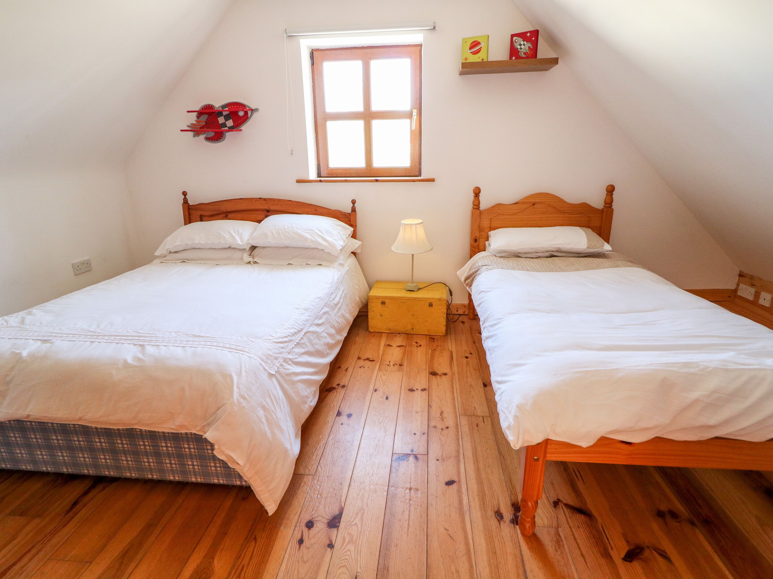 A bedroom with two beds, a window, a nightstand, and a shelf with books.