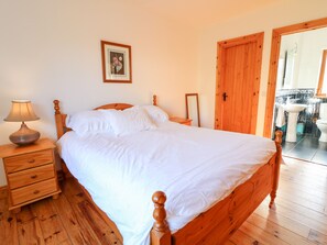 A neatly made bed with white linens, a wooden nightstand with a lamp, and a wooden door leading to a bathroom. - Crolly Home (Crolly)