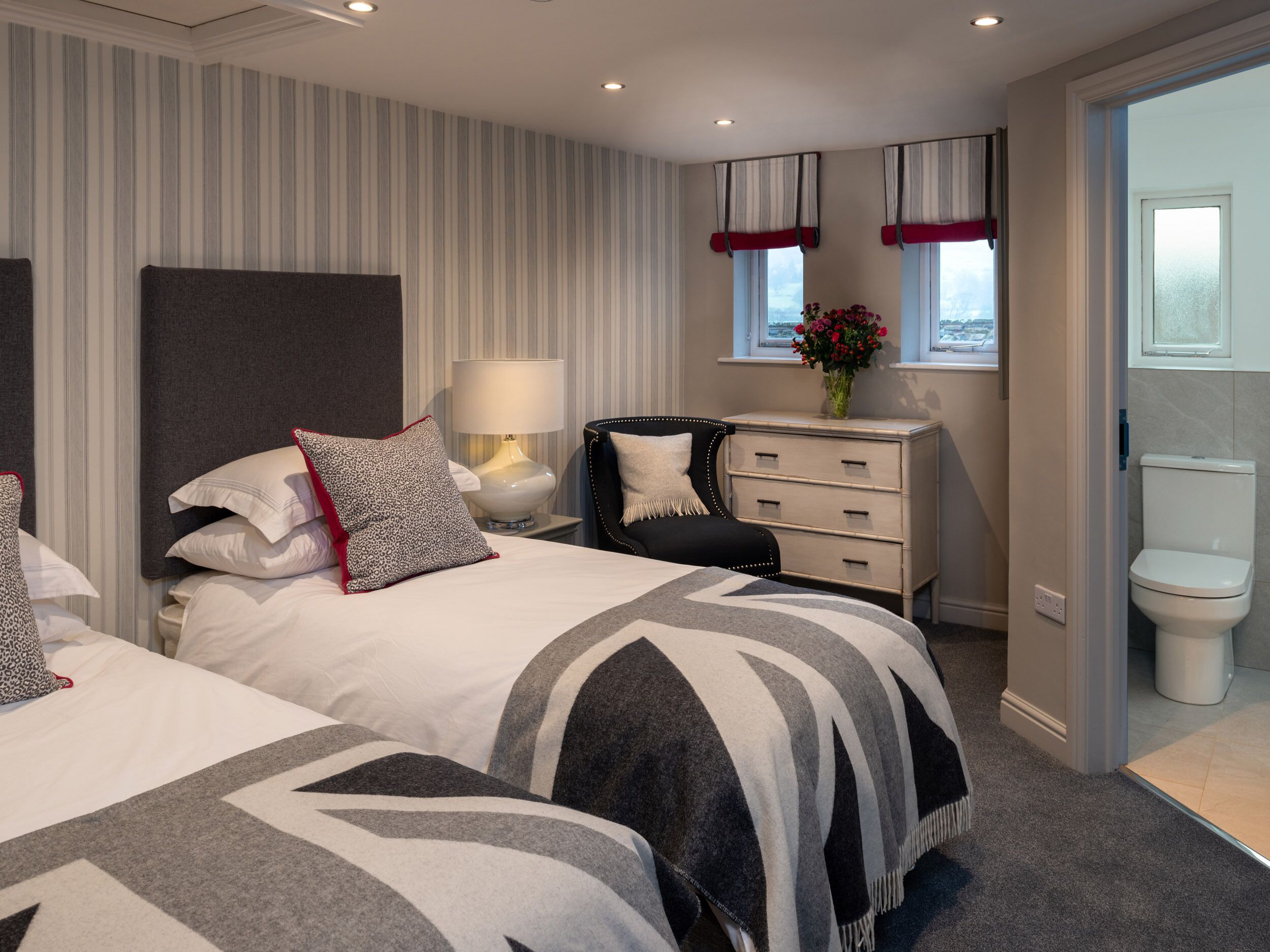 A hotel room with two beds, a bedside table with a lamp, a dresser, a window with curtains, and a toilet.