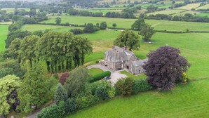 Une grande maison en pierre, chargée d’histoire, entourée de vastes champs verdoyants et d’arbres.