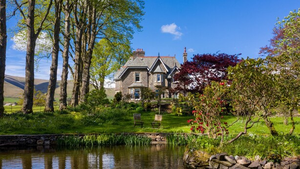 Une maison en pierre avec un étang devant, entourée d’arbres et d’une zone herbeuse.