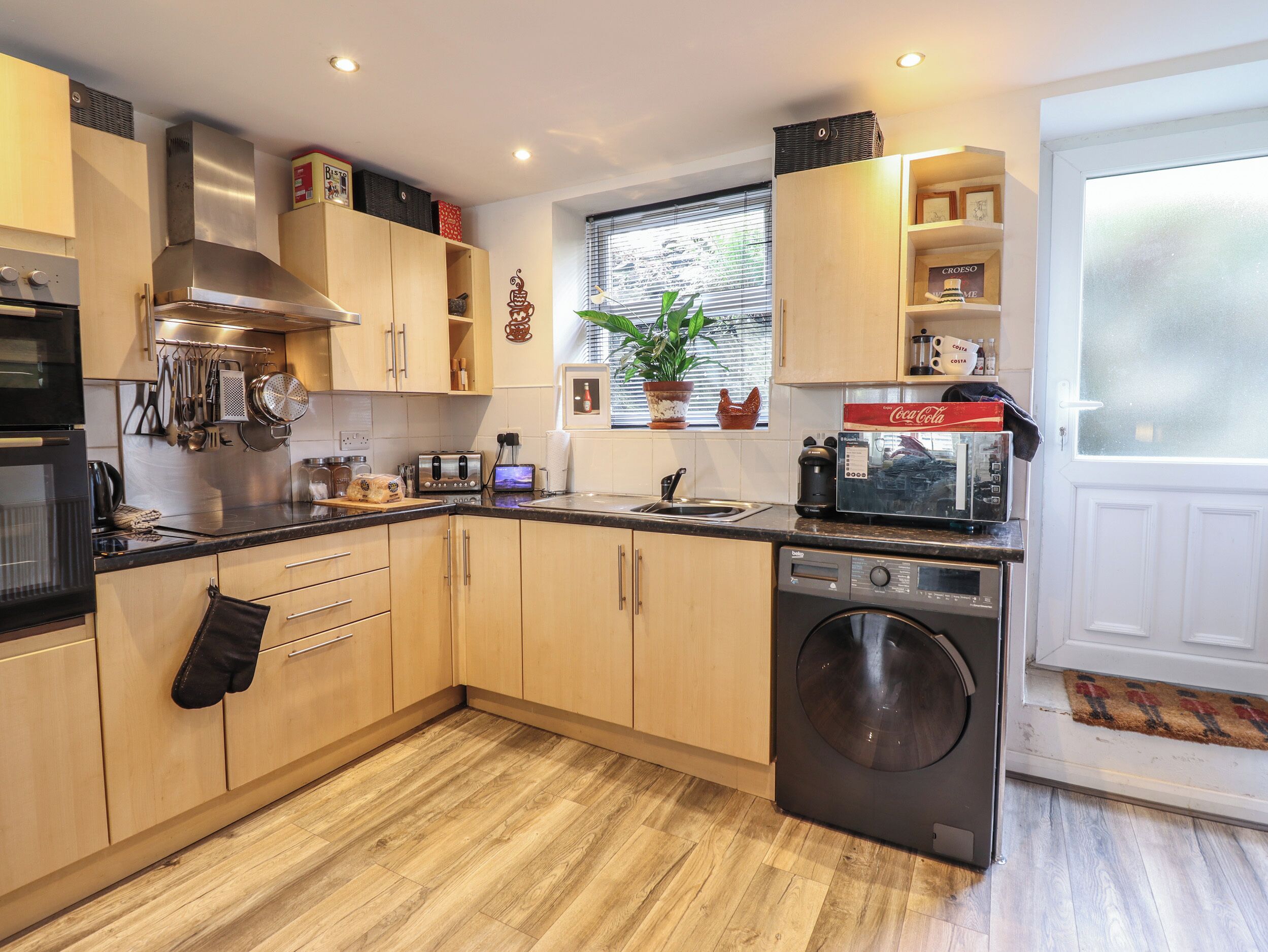 A modern kitchen with wooden cabinets, a built-in oven, a washing machine, and a window with blinds.