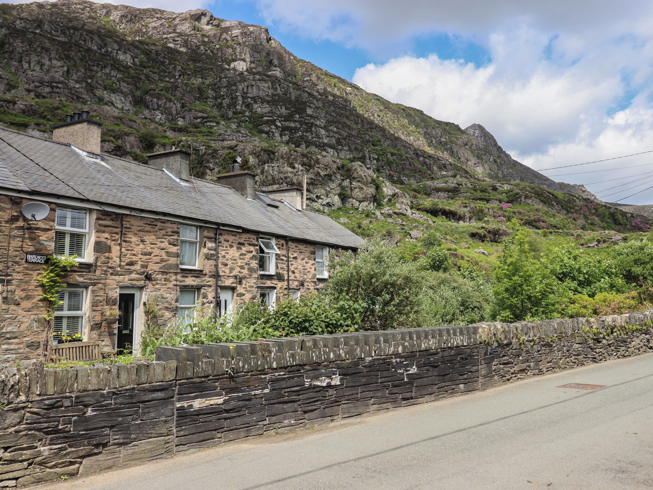 A stone house with a sloped roof, a stone wall in front, and a mountain in the background.
