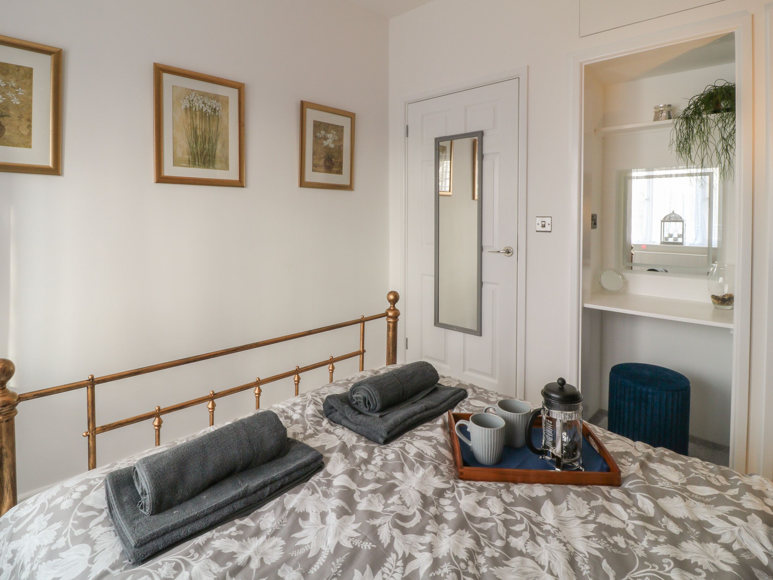 A neatly made bed with a floral bedspread, a bedside table with a tray holding a coffee pot, cups, and a book, and a mirror above the door.