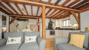Een ruime woonkamer met grijze banken en houten balken, een keuken met moderne apparatuur, en een trap naar een andere kamer.