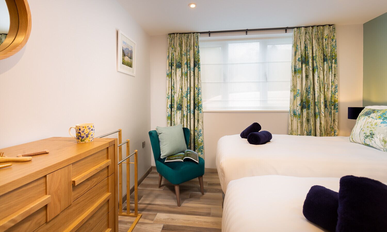 Ein Hotelzimmer mit einem Holzbüro, einem Türkissessel, einem Bett mit dunkelblauen Kissen und blumigen Vorhängen.