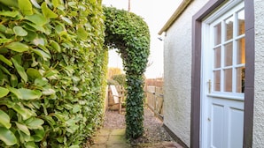 A narrow pathway leading to a white door, flanked by dense green foliage on one side and a stone wall on the other. - Rhum Cottage (Cupar)