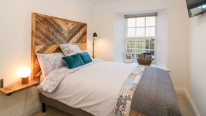 A bedroom with a bed, a nightstand with a lamp, a window, and a television on the wall. - Rhum Cottage (Cupar)