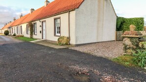 A row of traditional houses with red tiled roofs and white walls, a gravel driveway, and a stone wall with greenery. - Rhum Cottage (Cupar)