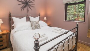 A bedroom with a metal bed frame, white bedding, a decorative wall piece, a window with a view of greenery, and a bedside table with a lamp. - Corner Cottage (Windermere)