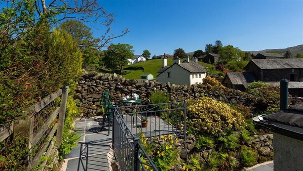 A stone wall with a metal gate, a small table with chairs, and a stone building in the background. - Corner Cottage (Windermere)