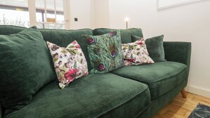 A green sofa with floral cushions in a room with a wooden floor and a framed picture on the wall. - Corner Cottage (Windermere)