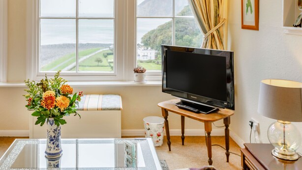 A room with a television on a wooden table, a vase with flowers on a glass table, and a view of the sea through large windows. - Clooneavin Apartment 2 (Lynmouth)