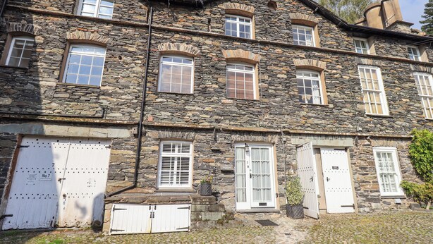 A stone building with multiple windows and doors, a cobblestone ground, and a small garden area. - The Mounting Steps (Ambleside)