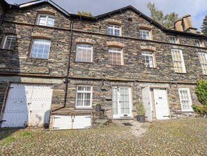 Um edifício de pedra com várias janelas e portas, chão de paralelepípedos e uma pequena área ajardinada.