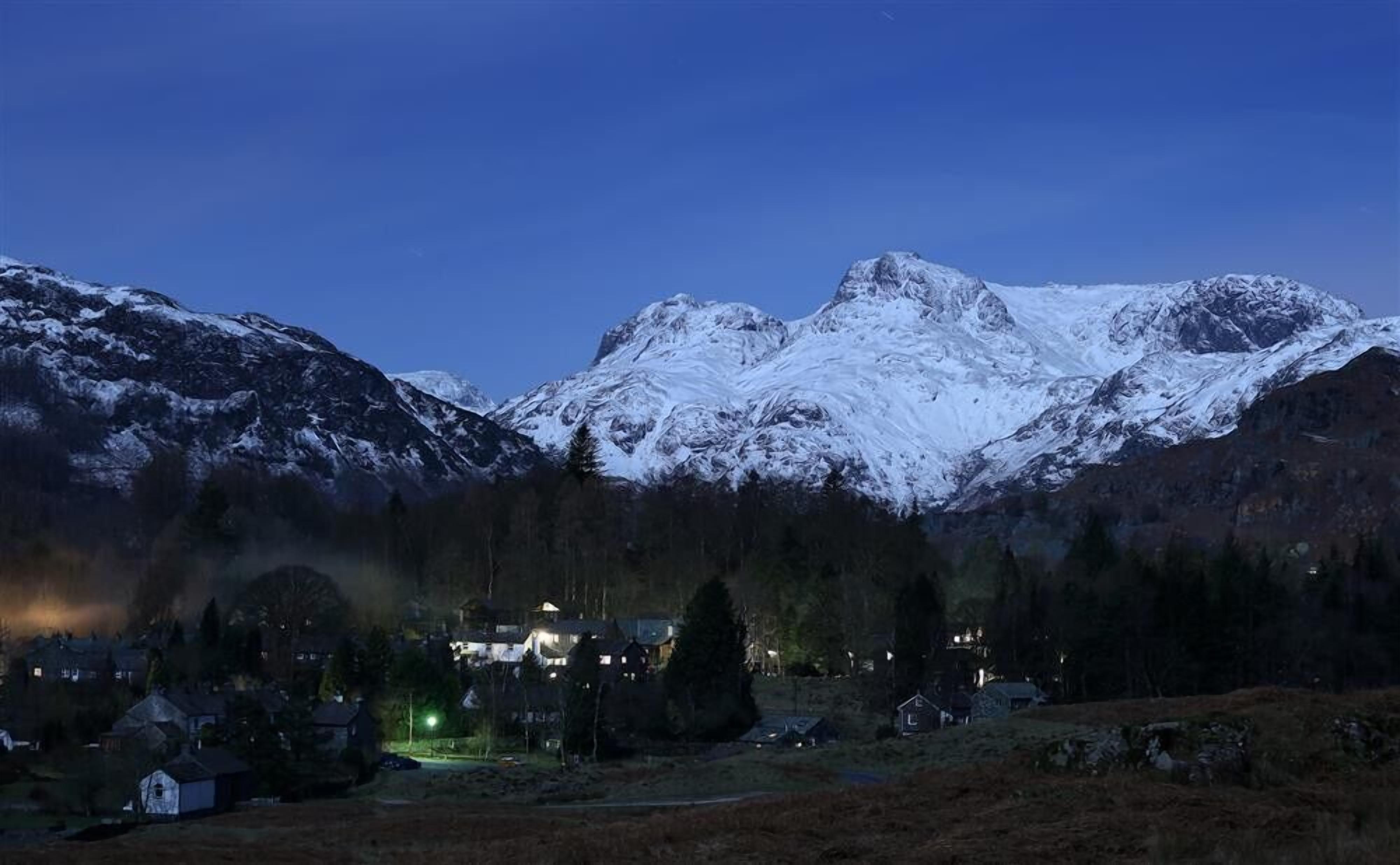 Un paisaje montañoso con picos nevados y un pequeño poblado con casas y una calle iluminada.