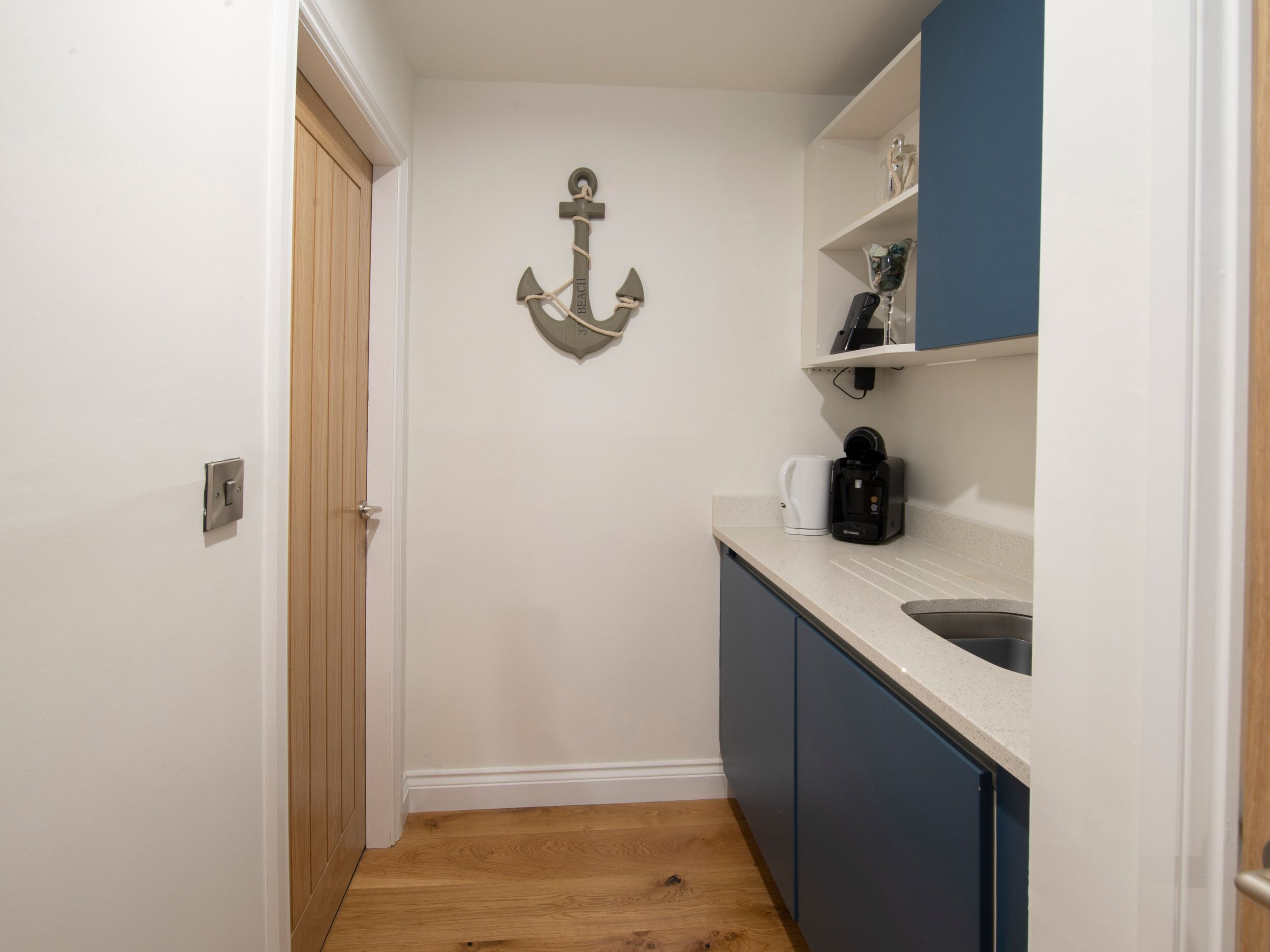 A small, narrow kitchen with a wooden floor, a white door, a shelf with decorative items, and a coffee maker.