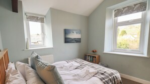 Une chambre à coucher avec un lit, des oreillers, un radiateur, une bibliothèque, un tableau et deux fenêtres avec des rideaux.