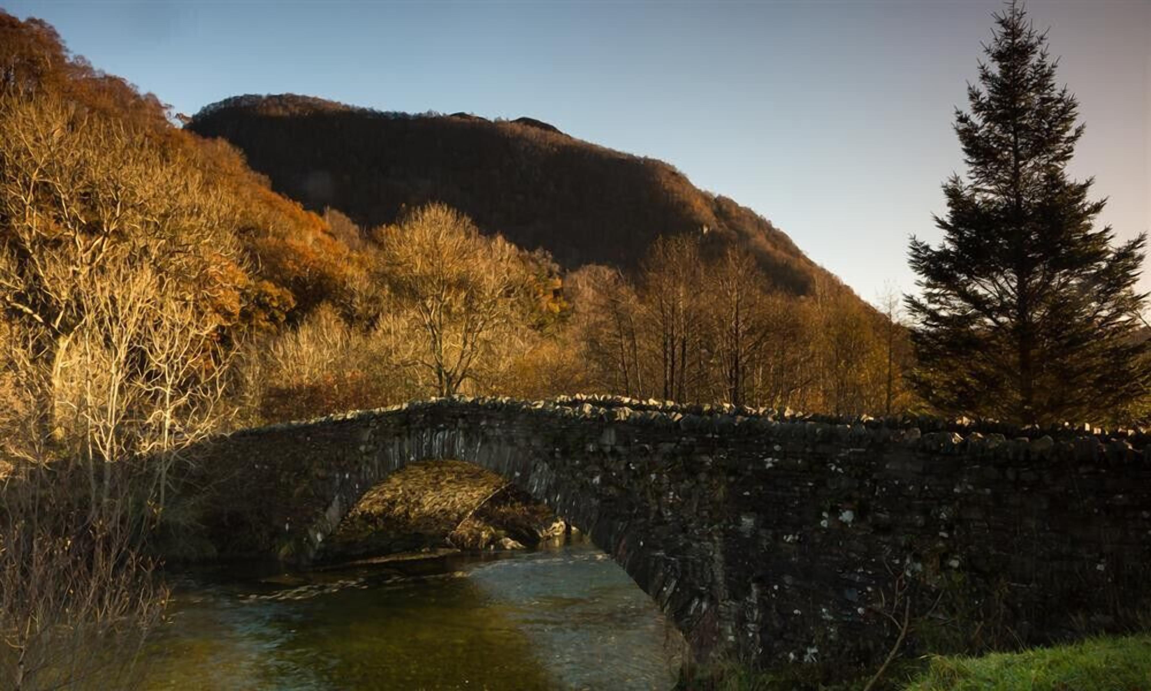 Un pont de pierre enjambant une rivière, avec une colline boisée en arrière-plan.