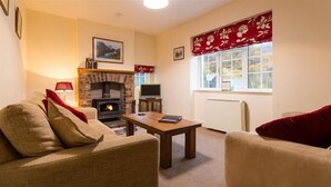 A cozy living room with a fireplace, a sofa, a table, and a window with a view of the outdoors. - Grange Fell (Keswick)