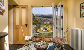 Un balcon accueillant avec une table préparée pour le petit-déjeuner, des chaises et une vue sur des collines verdoyantes.