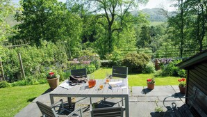 An outdoor patio with a table and chairs, surrounded by lush greenery and a view of hills. - Oakbank (Ambleside)