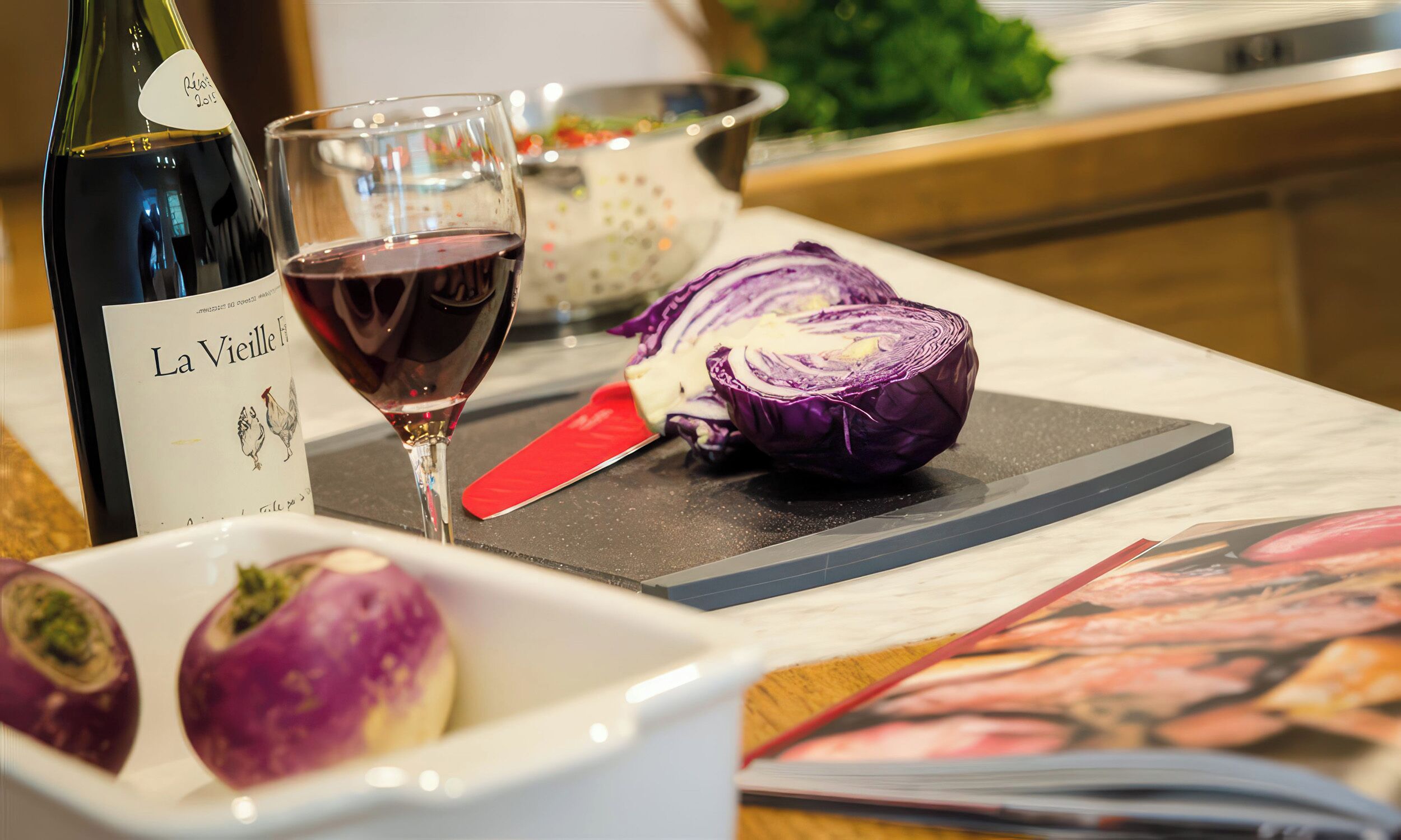 A bottle of wine, a glass of red wine, a halved red cabbage, and a white bowl with two purple vegetables on a kitchen counter.