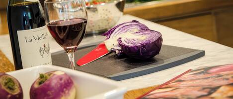 A bottle of wine, a glass of red wine, a halved red cabbage, and a white bowl with two purple vegetables on a kitchen counter.