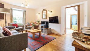 A living room with a sofa, armchair, coffee table, and a fireplace. There is a television mounted on the wall and a window with curtains. - Oakbank (Ambleside)