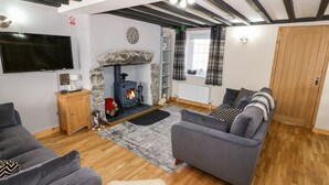 A cozy living room with a stone fireplace, grey sofas, a flat-screen TV, and a wooden ceiling. - Min-y-rhos (Pwllheli)