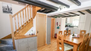 A dining area with a wooden table and chairs, a kitchen in the background, and a staircase leading to another level. - Min-y-rhos (Pwllheli)