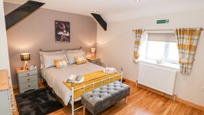 A bedroom with a bed, bedside tables, a bench, a radiator, a window with curtains, and a painting on the wall. - Min-y-rhos (Pwllheli)