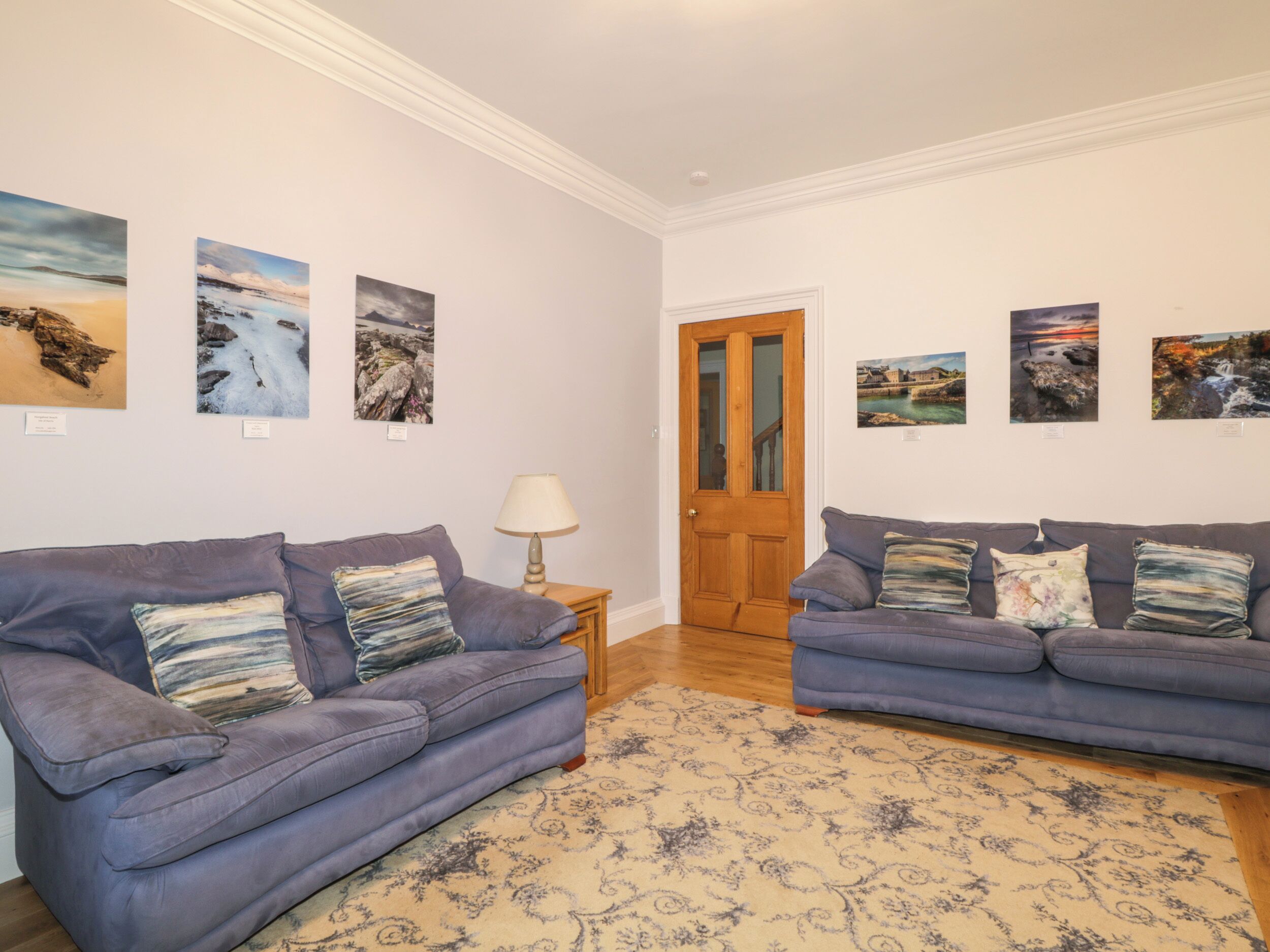 Een woonkamer met twee blauwe banken, een kamerbreed tapijt met dessin en verschillende landschapsschilderijen aan de wand.
