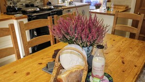 A wooden dining table with a wicker basket of bread, a vase with pink flowers, and a bottle of Black Isle Dairy Whole Milk.