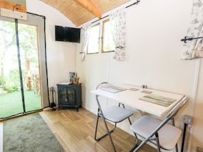 A room with a wooden ceiling, a television mounted on the wall, a table with chairs, and a window with patterned curtains. - Hugatree (King's Lynn)