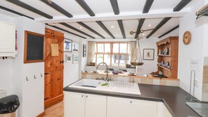 A modern kitchen with white cabinets, a black countertop, and a wooden door. There is a window with curtains, a sink with a dish rack, and a shelf with decorative items. - Cherry Tree Cottage at Satterthwaite (Ulverston)