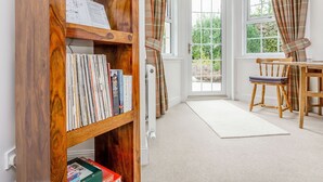 Una habitación con una estantería de madera llena de libros, un comedor con mesa y sillas de madera, y un amplio ventanal con cortinas.