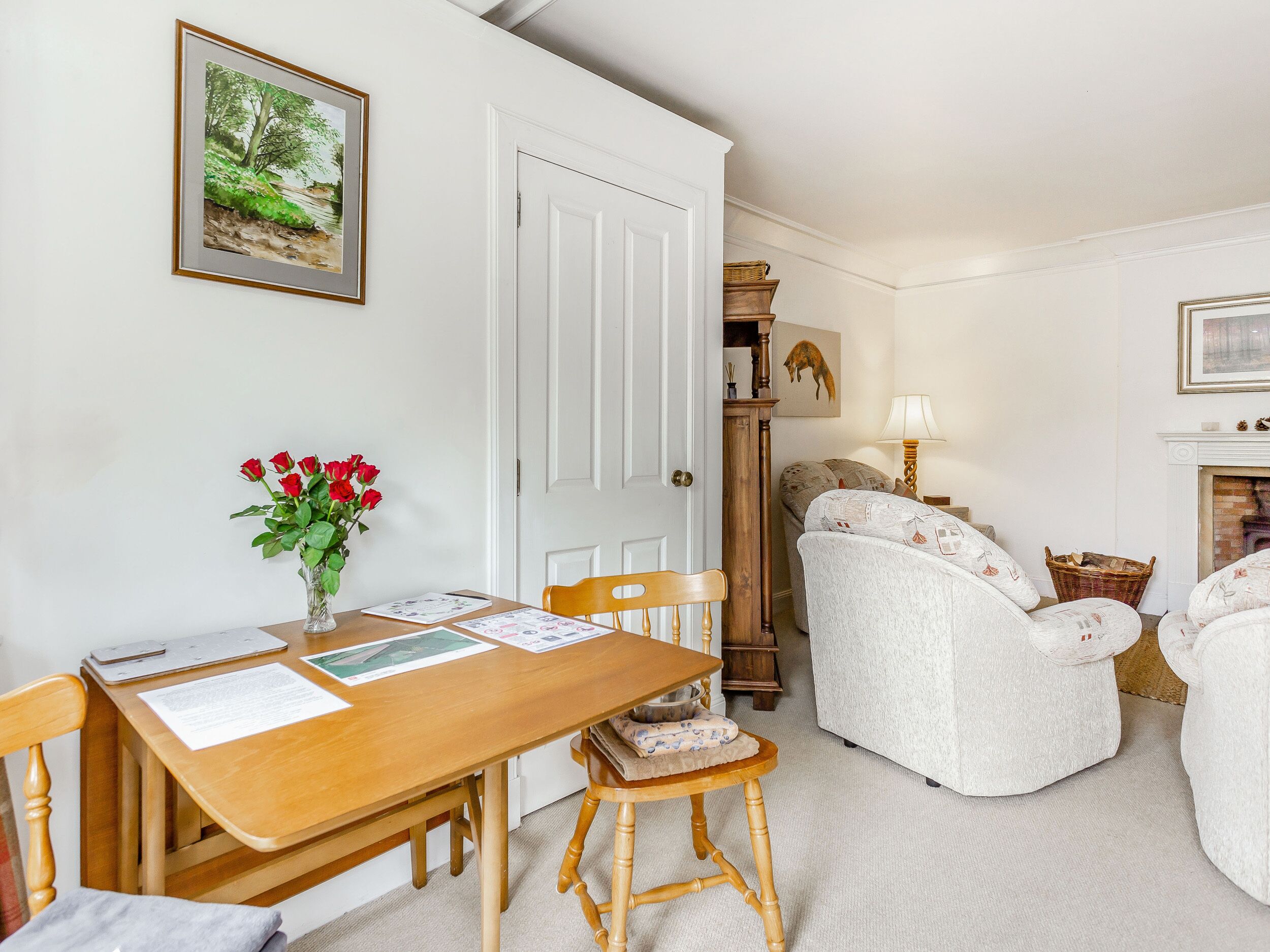 Een gezellige kamer met een houten tafel, stoelen, een vaas rode rozen, een open haard en een schilderij aan de muur.