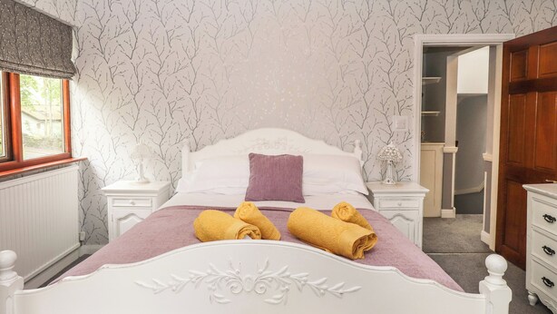 A neatly made bed with rolled towels, a white headboard with decorative carvings, a window with a view, and a wooden door leading to a closet.