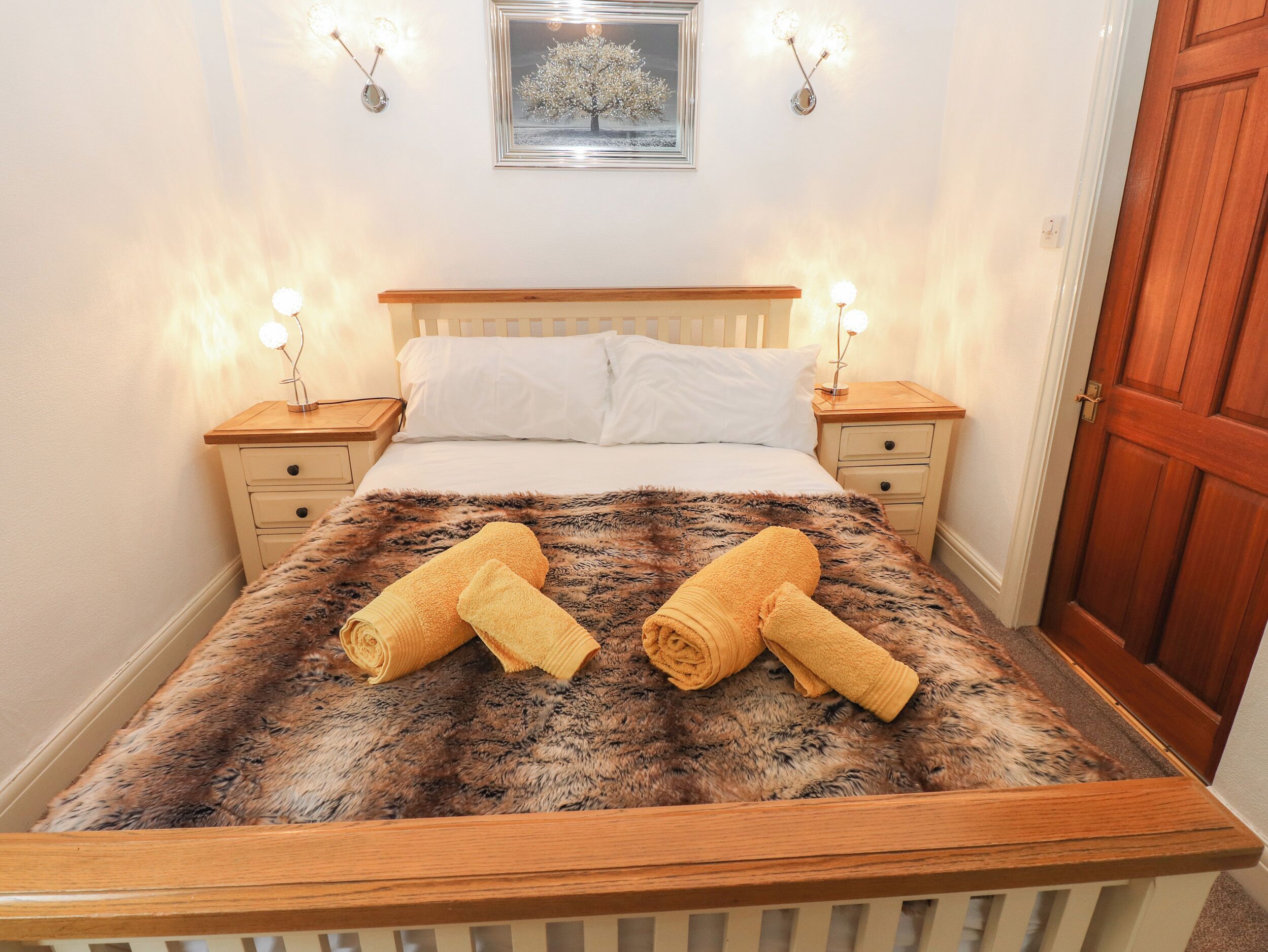 A neatly made bed with a fluffy blanket, two bedside tables with lamps, a framed picture on the wall, and a wooden door.