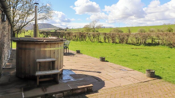 A wooden hot tub with steps and a table, surrounded by a stone patio and a grassy field with a vineyard in the background. - Home Farm House (Penrith)